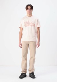 Hellrosa Baumwoll-T-Shirt mit einem Druck eines Krokodils, kombiniert mit beigen Jeans und schwarzen Sneakers. Das Model steht vor einem weißen Hintergrund.