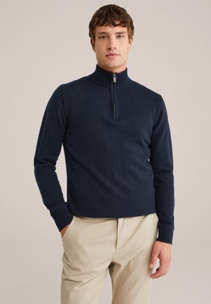 Pulover închisor navy cu fermoar, guler înalt, mâneci lungi, țesătură tricotată texturată și manșete ribelate, asortat cu pantaloni de culoare deschisă.