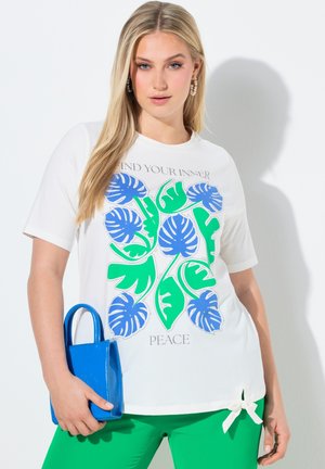 Kvinde iført hvid skjorte med blå og grønne bladprint og teksten "Find din indre fred," holder en lille blå håndtaske og er klædt i grønne bukser.