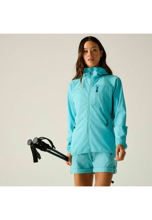 Donna che indossa una giacca azzurro chiaro e pantaloncini, che tiene in mano bastoncini da trekking neri su uno sfondo neutro.