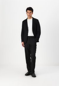 Črn blazer z dvema gumboma, v kombinaciji z belo majico in črnimi hlačami. Model nosi črne čevlje in stoji pred belo ozadjem.