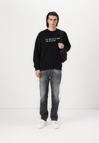 Sudadera negra con texto blanco, jeans grises y zapatillas blancas. Lleva una pequeña bolsa negra. Tejido suave y corte relajado.