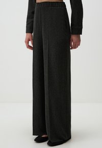 Pantalons larges à côtes noires avec taille élastique et poches latérales, présentés avec des chaussures plates noires. Texture douce et coupe décontractée.