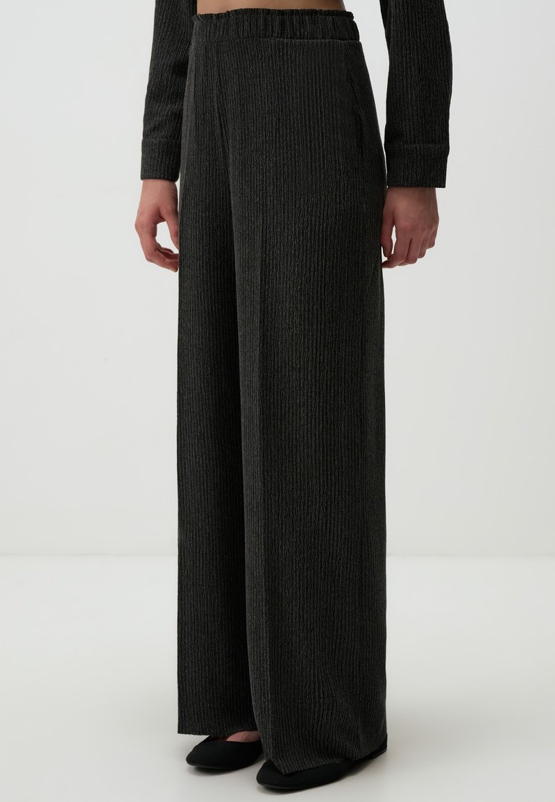 Pantalons larges à côtes noires avec taille élastique et poches latérales, présentés avec des chaussures plates noires. Texture douce et coupe décontractée.