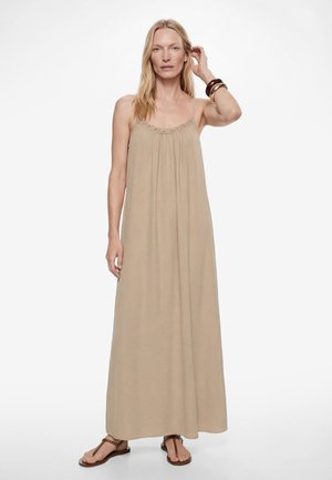 Femme portant une robe longue beige sans manches avec des bretelles spaghetti, des sandales marron et des bracelets, debout devant un fond clair.