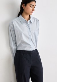 Camisa de botão de manga longa azul-claro com um bolso, combinada com calças de faixa elástica azul-marinho, contra um fundo neutro.