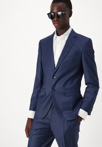 Navy blue suit with a textured fabric, slim fit design, notch lapels, two buttons, paired with a white shirt and black sunglasses.