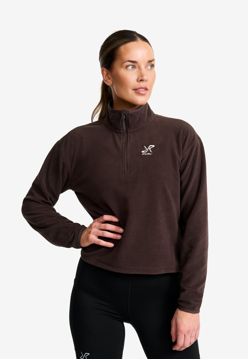 Frau mit braunem Fleece-Pullover mit Viertelzip und schwarzen Leggings, die mit einer Hand auf der Hüfte stehend zur Seite schaut, vor einem weißen Hintergrund.