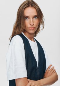 Jeansvest met diepe V-hals over een wit T-shirt met korte mouwen. Donkerblauwe stof met lichte stiksels, gladde textuur en getailleerde silhouet.