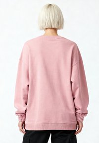 Personne aux cheveux blonds courts portant un sweat-shirt rose ample, vue de dos sur un fond blanc uni.