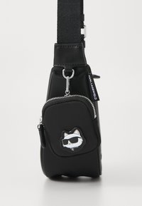 Bolso bandolera negro con acabado mate, que presenta un bolsillo con cremallera en la parte frontal y un parche con diseño de gato. Correa ajustable y detalles de hardware en color plateado.