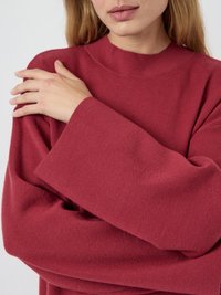 Frau in einem langen roten Pullover, die mit ihrer rechten Hand ihre linke Schulter gegen einen schlichten Hintergrund hält.