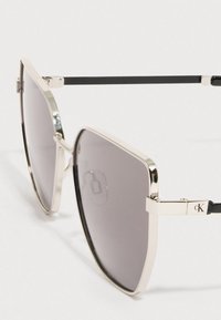 Prostokątne okulary przeciwsłoneczne z ciemnoszarymi soczewkami, srebrną metalową oprawą i czarnymi zausznikami. Po bokach znajduje się subtelne logo.