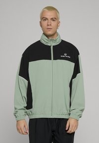 Lys grønn og svart jakke med glidelås, høy krage, elastisk midjebånd, logo på brystet og kontrasterende paneldesign. Glatt, lett stoff.