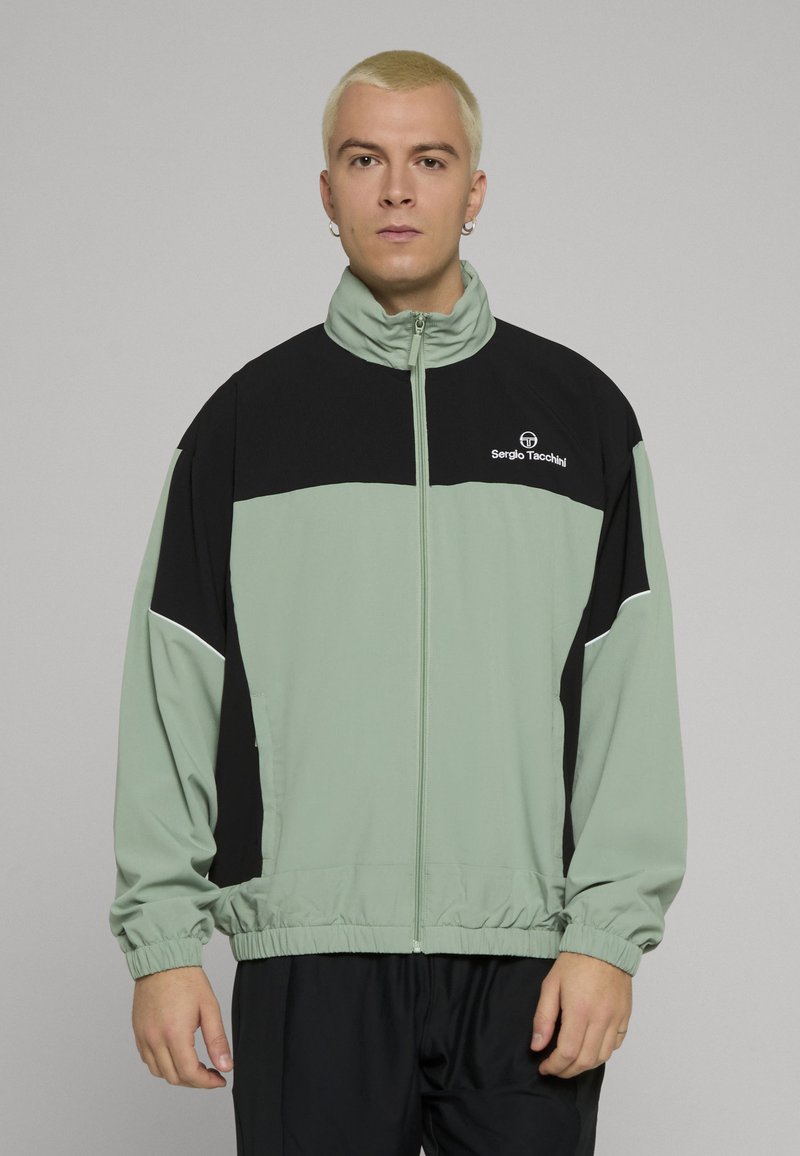 Lys grønn og svart jakke med glidelås, høy krage, elastisk midjebånd, logo på brystet og kontrasterende paneldesign. Glatt, lett stoff.