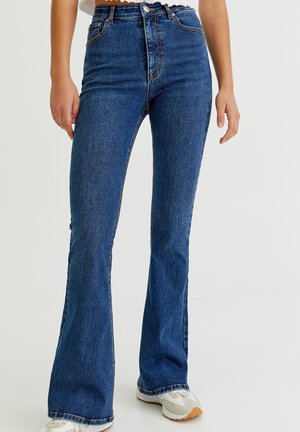 Femme portant un jean évasé taille haute bleu foncé et des baskets blanches, debout devant un fond clair uni.