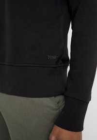 Sudadera negra en mezcla de algodón, con puños y dobladillo acanalados, y un sutil logo en relieve "DRYKORN" en la parte inferior izquierda. Combinada con pantalones verde oliva.