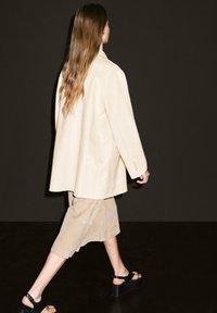 Beiger oversized Blazer, kombiniert mit einem beigen Wildleder-Rock und schwarzen Plateausandalen, vor einem dunklen Hintergrund. Glatte Textur, lässige Passform.