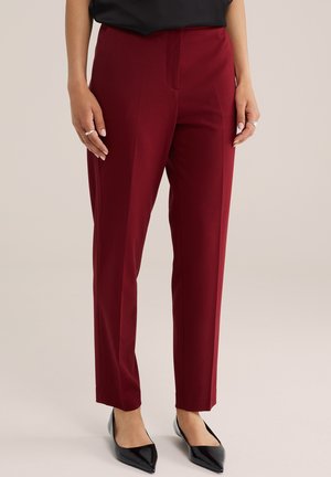 Femme portant un pantalon rouge foncé sur mesure et des ballerines noires pointues, debout devant un fond neutre.