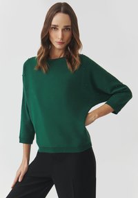 Pullover verde in maglia con scollatura ampia, maniche a tre quarti e dettagli in perline turchesi. Tessuto morbido e vestibilità comoda. Abbinato a pantaloni neri.