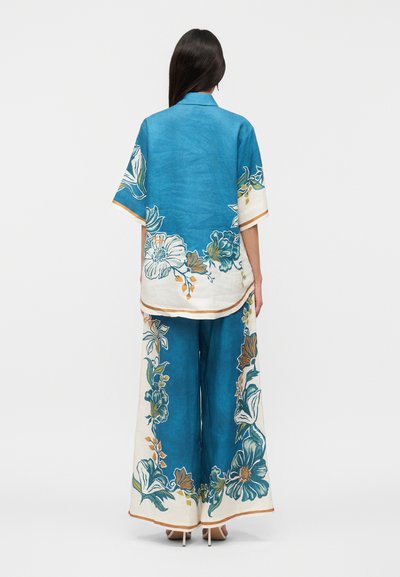 Γυναίκα που φοράει μπλε και λευκό φαρδιά παντελόνια με λουλουδάτο σχέδιο και αντίστοιχο άνετο κοντομάνικο τοπ, όρθια με την πλάτη στραμμένη.