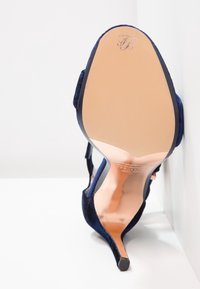 Mörkblå mocka högklackad sandal med tunna remmar, spetsig tå och en slät rosaguld inläggssula med en diskret logotyp på klacken.