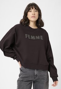 Mladá žena s tmavými vlasmi po pleciach oblečen�á v čiernej mikine s nápisom "FEMME" trblietavými písmenami a vyblednutými čiernymi džínsami.