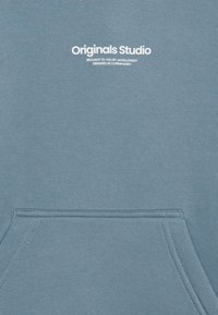 Svetlo modrý sveter s klokánkovou vreckom, vyrobený z jemnej bavlny. Na prednej strane obsahuje biely potlačený nápis "Originals Studio".