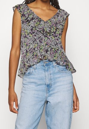 Kobieta w bezrękawnym, kwiecistym topie z falbankami, ozdobionym czarnymi, fioletowymi i żółtymi kwiatami, do tego wysokie, jasnoniebieskie dżinsy.