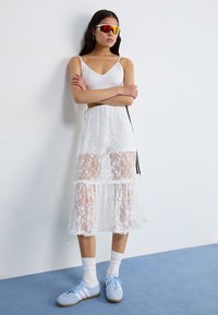 White lace maxi skirt with sheer sections and ruffled hem, paired with a fitted white top and light blue sneakers, featuring gray stripes.