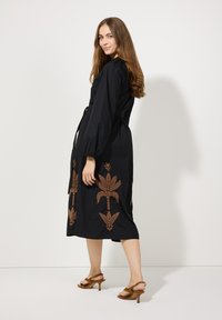 Schwarzes Kleid mit langen Puffärmeln, taillierter Taille und aufwendiger brauner floralem Stickerei am Saum. Kombiniert mit braunen Absatzsandalen.