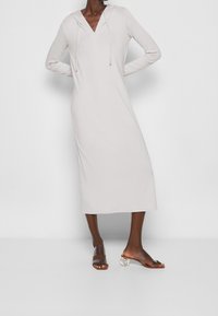 Lång, ljusgrå klänning med huva, långa ärmar och V-ringning. Slät textur med justerbara dragsnören. Bruna sandaler med släta, genomskinliga klackar.