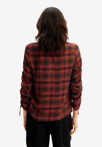 Donna con capelli scuri di lunghezza fino alle spalle, che indossa una camicia a maniche lunghe a quadri rossi e neri e pantaloni neri, vista da dietro.