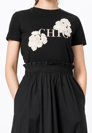 Mujer con camiseta negra de manga corta con bordado floral blanco y texto "CHIC", combinada con falda negra de talle alto con cintura fruncida.