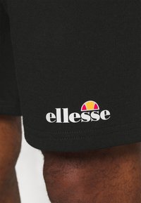 Mustat puuvillashortsit, joissa on valkoinen "ellesse"-logo ja monivärinen graafinen yksityiskohta sen yläpuolella, joka esittää puolipallon muotoista suunnittelua.