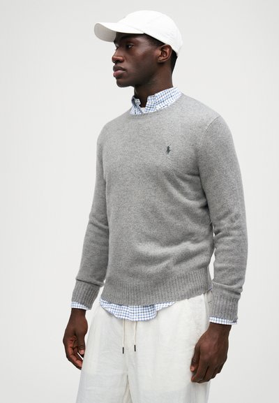 Suéter cinza em tecido suave, gola redonda, punhos e bainha canelados, usado sobre uma camisa xadrez azul, combinado com calças de cor clara.