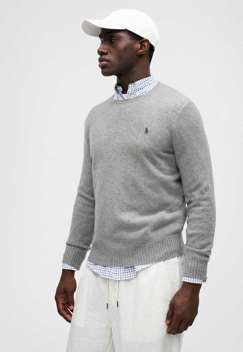 Suéter gris de tejido suave, con cuello redondo, puños y dobladillo acanalados, llevado sobre una camisa a cuadros azul, combinado con pantalones de color claro.