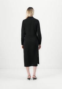 Black long-sleeve dress with a straight cut, side slits, and buttoned cuffs, made of lightweight fabric; worn with strappy gray heels.
