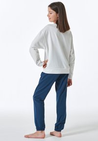 Weißes Langarmoberteil mit gerippten Bündchen, kombiniert mit blauen Jogginghosen mit elastischen Knöcheln. Das Model hat glattes Haar und posiert von hinten.