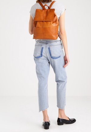 Vrouw draagt lichtblauwe spijkerbroek, zwarte loafers en een bruine leren rugzak, gezien van achteren tegen een effen witte achtergrond.