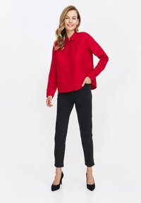 Czerwona koszula z długim rękawem, z kołnierzykiem, z zapięciem na guziki oraz czarne dżinsy slim-fit. Modelka ma na sobie czarne szpilki. Gładkie białe tło.