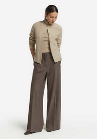 Beige strikket cardigan med ståkrave, parret med en lys beige ribbet sweater og brede brune bukser. Glat tekstur, skræddersyet pasform.