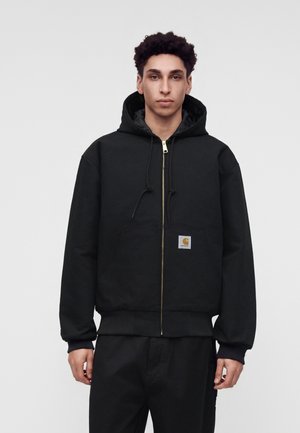 Un jeune homme porte un sweat à capuche noir avec une fermeture éclair et un petit patch logo rectangulaire sur la poche avant, debout devant un fond clair uni.