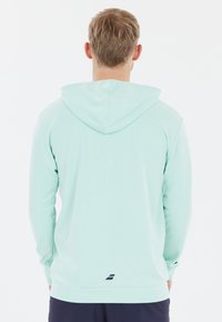 Lys grøn langærmet hoodie med hætte, med en lige kant og et lille navy logo nær nederste venstre side. Blød tekstur.