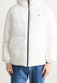 Vit pufferjacka med frontdragkedja, ribbade ärmslut och huva. Har horisontella sömmar och en liten logotyp på bröstet.