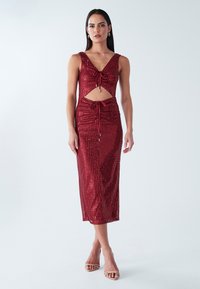 Sarkana fliterētu kleita ar piegulošu siluetu, V veida izgriezumu, vidēja garuma griezumu, izgriezuma detaļu viduklī un aukliņu akcentiem.