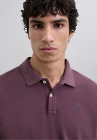 Polo de color marrón burdeos hecho de algodón, con un cuello clásico, dos botones y un pequeño logotipo de calavera en color marino en el lado izquierdo del pecho. Textura suave.