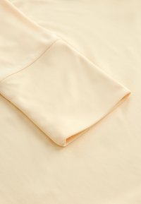 Tissu pêche clair avec une texture lisse ; présente un détail de manche repliée mettant en valeur des lignes épurées et une finition douce.