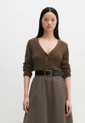 Vrouw die een bruine gebreide cardigan met V-hals en opgerolde mouwen draagt, ingestopt in een hoge taille taupekleurige rok met een zwarte riem en gouden gesp.