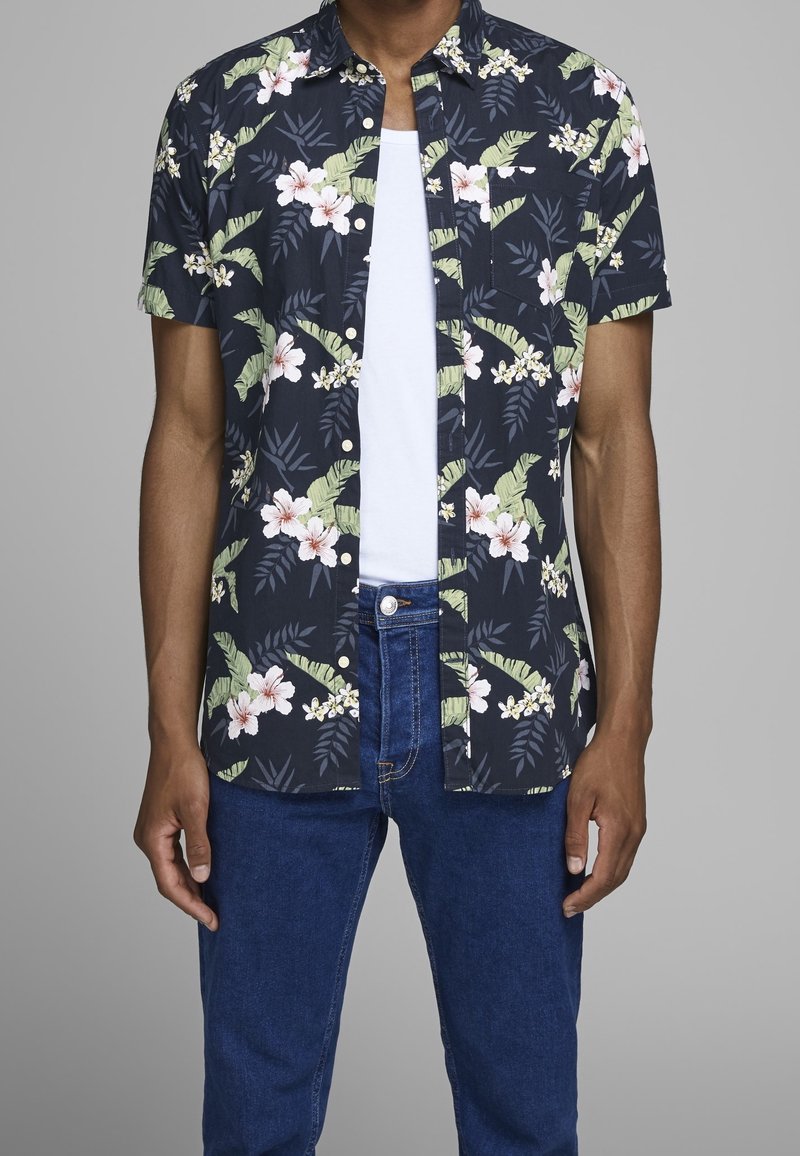 Uomo che indossa una camicia a maniche corte con fantasia floreale blu navy sopra una maglietta bianca e jeans blu, in piedi contro uno sfondo grigio semplice.